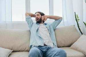 uma homem senta em a sofá bocejando e alongamento depois de uma sesta, falta do dormir e fadiga a partir de trabalhos e impróprio diariamente rotina. foto
