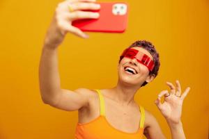 mulher sorrisos e mostra dela língua dentro incomum óculos milenar leva selfies em dela telefone dentro roupa de esporte contra a laranja estúdio pano de fundo, livre espaço foto