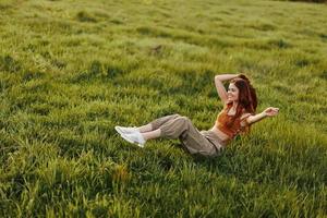 uma jovem, feliz mulher jogando verão jogos e queda em a verde Relva dentro a parque dentro a configuração Sol e sorridente. em repouso e jogando dentro a fresco aberto ar foto