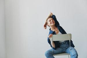 uma jovem mulher sentado dentro uma cadeira às casa sorridente com dentes com uma curto corte de cabelo dentro jeans e uma jeans camisa em uma branco fundo. menina natural poses com não filtros foto