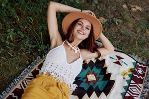 uma mulher dentro ecológico roupas dentro uma hippie Veja é deitado em uma colori xadrez sorridente e olhando às a outono pôr do sol dentro natureza dentro a parque. estilo de vida em uma viagem do harmonia com a mundo foto