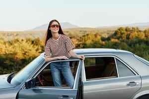 uma jovem mulher motorista parece Fora do a carro às a outono panorama e sorrisos satisfatoriamente foto