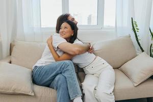 uma homem e uma mulher estão sentado em a sofá às casa vestindo branco Camisetas e abraçando cada de outros com sorri. família vida estilos de vida do jovem casados foto