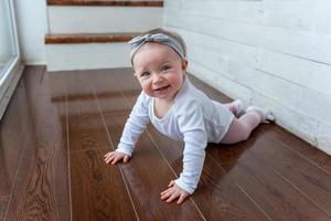 garotinha rastejante de um ano de idade sentada no chão na sala de estar de luz brilhante perto da janela sorrindo e rindo foto