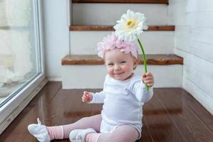 pequeno bebê menina vestindo Primavera guirlanda localização em chão dentro brilhante luz vivo quarto perto janela e jogando com gerbera flores foto