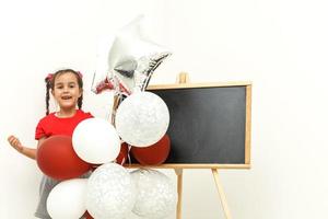 feliz brilhante imagem do fofa alegre pequeno menina dentro tule saia sentado em presente com balões isolado em branco fundo. surpreendente encantador aniversário elegante criança olhando para Câmera foto