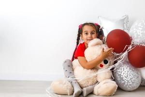 fofa criança pequeno menina posando com vermelho coração em forma balões e uma Urso de pelúcia Urso isolado em branco. crianças moda foto