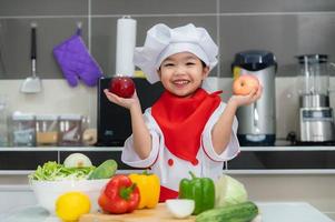 fofa ásia menina vestem chefe de cozinha uniforme com uma muitos do vegetal em a mesa dentro a cozinha quarto, fazer Comida para comer jantar, engraçado Tempo para crianças foto