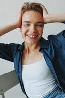 uma jovem mulher sentado dentro uma cadeira às casa sorridente com dentes com uma curto corte de cabelo dentro jeans e uma jeans camisa em uma branco fundo. menina natural poses com não filtros foto