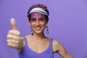retrato do uma desportivo moda mulher posando sorridente com dentes e apontando uma dedo às a Câmera dentro uma roxa ioga fato de treino e uma transparente boné em uma roxa monocromático fundo foto