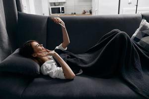 uma mulher mentiras em uma sofá dentro a apartamento com uma Móvel telefone dentro dela mão, coberto de uma cinzento cobertor foto