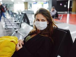 mulher vestindo uma médico mascarar sentado às a aeroporto esperando para uma voar amarelo mochila foto