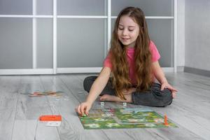 fofa pequeno menina com fluindo cabelo senta em a chão às casa e goza jogando uma borda jogos foto