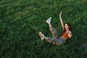 uma jovem mulher é jogando dentro a parque com nos dentro a Relva e cai para a terra sorridente alegremente dentro a luz solar. em repouso dentro natureza, harmonia com a corpo foto