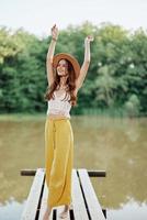 uma jovem mulher dentro uma hippie Veja e vestido ecológico viaja dentro natureza de a lago vestindo uma chapéu e amarelo calça dentro a outono pôr do sol foto