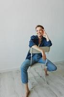 uma jovem mulher sentado dentro uma cadeira às casa sorridente com dentes com uma curto corte de cabelo dentro jeans e uma jeans camisa em uma branco fundo. menina natural poses com não filtros foto