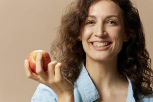 fechar-se retrato do alegre encaracolado fofa senhora dentro jeans casual camisa com maçã perto bochecha posando isolado em bege pastel fundo. saudável Comida. natural ecológico produtos conceito. cópia de espaço foto