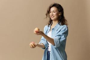 feliz sorridente focado encaracolado lindo fêmea dentro jeans casual camisa malabarismo com maçã posando isolado em sobre bege pastel fundo. saudável Comida. natural ecológico produtos conceito. cópia de espaço foto