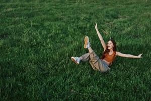 uma jovem mulher jogando jogos dentro a parque em a verde Relva espalhando dela braços e pernas dentro diferente instruções queda e sorridente dentro a verão luz solar foto