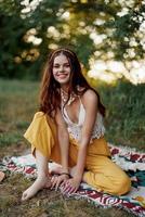 uma jovem lindo hippie mulher senta em a terra dentro a outono dentro eco roupas dentro a pôr do sol luz e sorrisos Atenciosamente para a Câmera foto