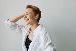 jovem mulher adolescente ouvindo para música com infértil fones de ouvido e dançando lar, sorrindo com dentes com uma curto corte de cabelo dentro uma branco camisa em uma branco fundo. menina natural poses com não filtros foto