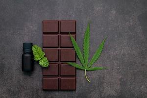 folha de cannabis com chocolate amargo, folhas de plantas e utensílios de madeira em um fundo escuro de concreto foto