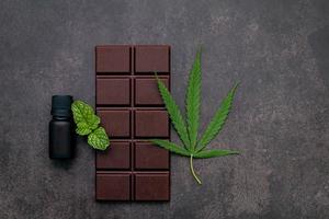 folha de cannabis com chocolate amargo, folhas de plantas e utensílios de madeira em um fundo escuro de concreto