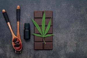 folha de cannabis com chocolate amargo, folhas de plantas e utensílios de madeira em um fundo escuro de concreto foto