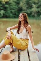 uma hippie mulher senta com dela chapéu em uma ponte de uma lago com dela mãos acima dentro a ar em uma natureza viagem e sorridente alegremente dentro roupas ecológicas. relaxado estilo de vida foto