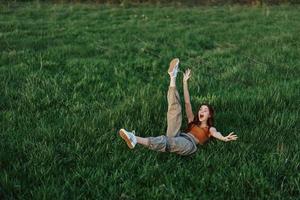 uma jovem mulher jogando jogos dentro a parque em a verde Relva espalhando dela braços e pernas dentro diferente instruções queda e sorridente dentro a verão luz solar foto