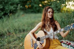 menina hippie mulher jogando guitarra dentro ecológico roupas sentado em a terra lado de fora dentro natureza dentro a outono assistindo a pôr do sol foto