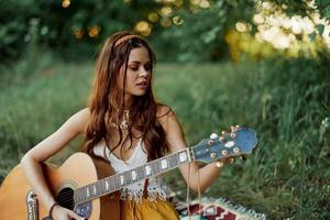 menina hippie mulher jogando guitarra dentro ecológico roupas sentado em a terra lado de fora dentro natureza dentro a outono assistindo a pôr do sol foto