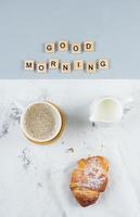 Boa manhã café da manhã mínimo conceito. copo do café, croissant e texto Boa manhã. plano deitar foto