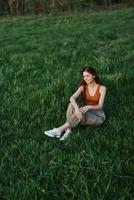 trabalhador autonomo mulher desfrutando a ao ar livre sentado dentro a parque em a verde Relva dentro casual roupas com grandes vermelho cabelo aceso de a brilhante verão Sol sem mosquitos foto