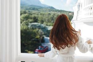 bonita mulher branco roupão admirar a Visão a partir de a janela inalterado foto