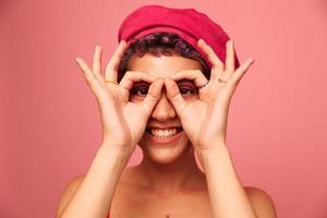 jovem Atlético mulher com uma curto corte de cabelo e roxa cabelo dentro uma vermelho topo e uma Rosa chapéu com a Atlético figura sorrisos e caretas mostrando sinais com dela mãos olhando às a Câmera em uma Rosa fundo foto