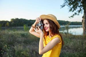 mulher fotógrafo com Câmera olhando para dentro a Câmera lente sorrir vermelho lábios chapéu natureza foto