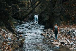 mulher dentro a montanhas perto a rio com uma mochila em dela costas e uma floresta dentro a fundo foto