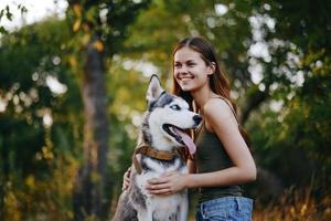 uma mulher com uma rouco procriar cachorro sorrisos e carinhosamente golpes dela Amado cachorro enquanto caminhando dentro natureza dentro a parque dentro outono contra a pano de fundo do pôr do sol foto