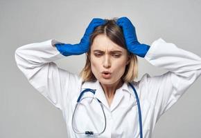 emocional mulher dentro azul médico luvas toques dela cabeça com dela mãos em uma luz fundo foto