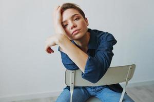 uma jovem mulher sentado dentro uma cadeira às casa sorridente com dentes com uma curto corte de cabelo dentro jeans e uma jeans camisa em uma branco fundo. menina natural poses com não filtros foto