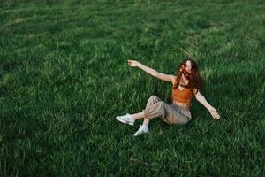 jovem mulher jogando jogos dentro a parque em a verde Relva espalhando dela braços e pernas dentro diferente instruções queda e sorridente dentro a luz solar do verão, uma estilo de vida do vida e juventude foto