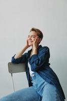 uma jovem mulher sentado dentro uma cadeira às casa sorridente com dentes com uma curto corte de cabelo dentro jeans e uma jeans camisa em uma branco fundo. menina natural poses com não filtros foto