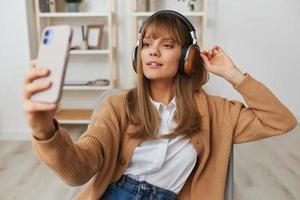 alegre jovem Loiras senhora dentro caloroso suéter dentro fones de ouvido ouço fav canções fazendo selfie vídeo ligar sentado dentro poltrona às moderno casa interior. música Tempo relaxante legal lista de reprodução conceito. cópia de espaço foto