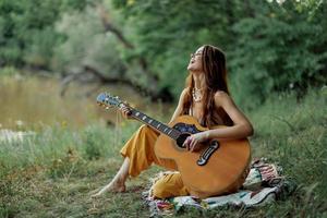 jovem hippie mulher sentado ao ar livre em uma xadrez e jogando dela guitarra em a margem do rio para dentro a pôr do sol foto