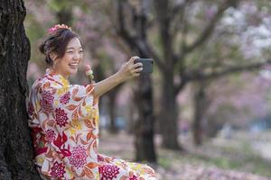 japonês mulher dentro tradicional quimono levando selfie hanami dango gesto enquanto caminhando dentro a parque às cereja Flor árvore durante Primavera sakura festival conceito foto