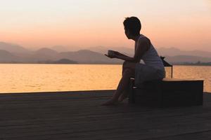 silhueta mulher segurando copo café sentado de a lago Assistir nascer do sol. foto