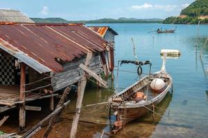 tailandês pescaria grandes barco Próximo para a de pescador cabana em pé em palafitas dentro a água. oxidado barraco teto. uma pescador derrama água a partir de uma barco. grande bóia dentro a barco. foto