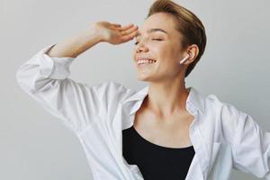 jovem mulher adolescente ouvindo para música com infértil fones de ouvido e dançando lar, sorrindo com dentes com uma curto corte de cabelo dentro uma branco camisa em uma branco fundo. menina natural poses com não filtros foto