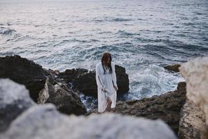 uma mulher dentro uma branco vestir carrinhos em rochoso pedras de a oceano inalterado foto